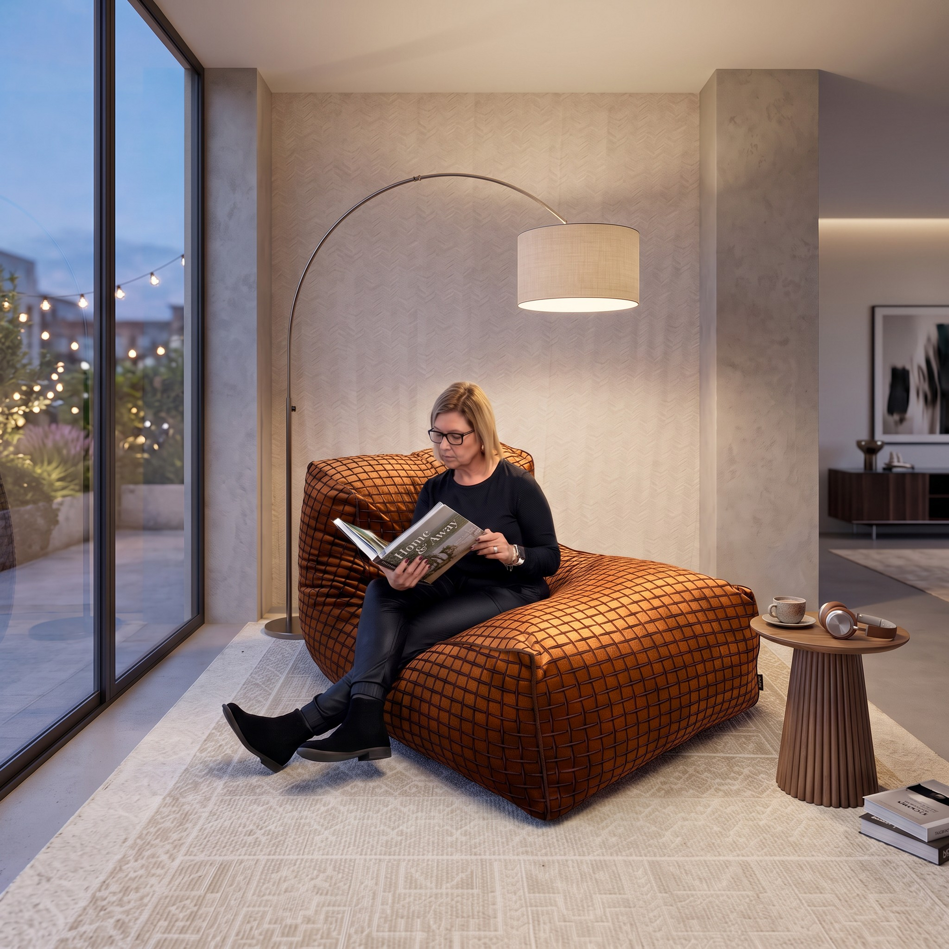 A woman sits on a large orange-brown checkered lounge chair in a modern living room, reading a magazine. The room features a tall arched floor lamp, a small side table with coffee cups, and a large window looking out onto a garden at dusk.