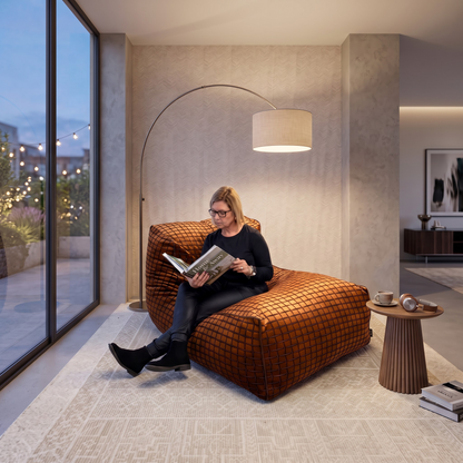 A woman sits on a large orange-brown checkered lounge chair in a modern living room, reading a magazine. The room features a tall arched floor lamp, a small side table with coffee cups, and a large window looking out onto a garden at dusk.