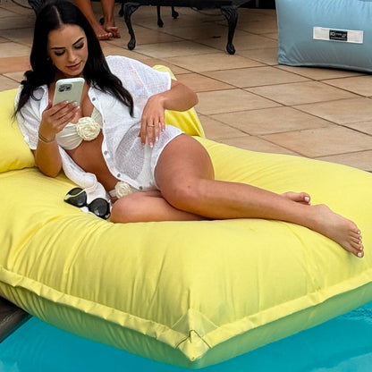 Woman lying on a yellow floating beanbag lounger by a pool, using a phone.